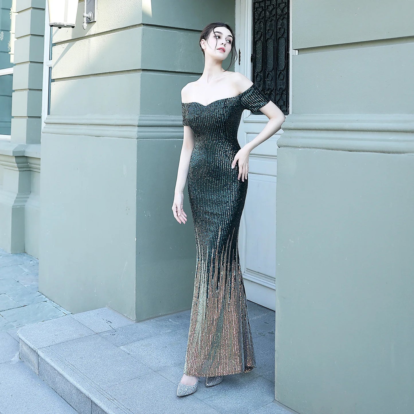 Woman in a black off-shoulder evening gown standing against a light gray building.