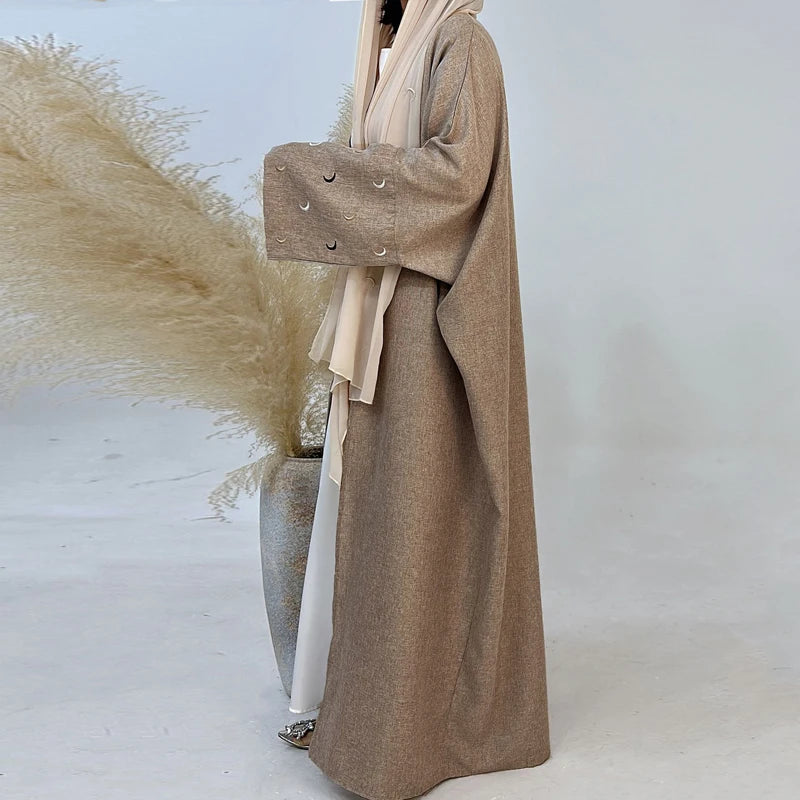 Person wearing a long brown coat standing next to a vase with dried plants on a light background