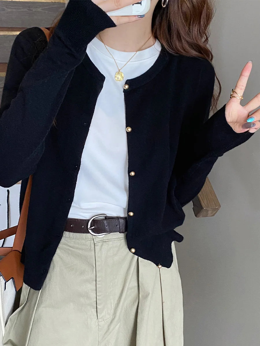 Person wearing a black cardigan over a white shirt with beige pants, standing against a neutral background.
