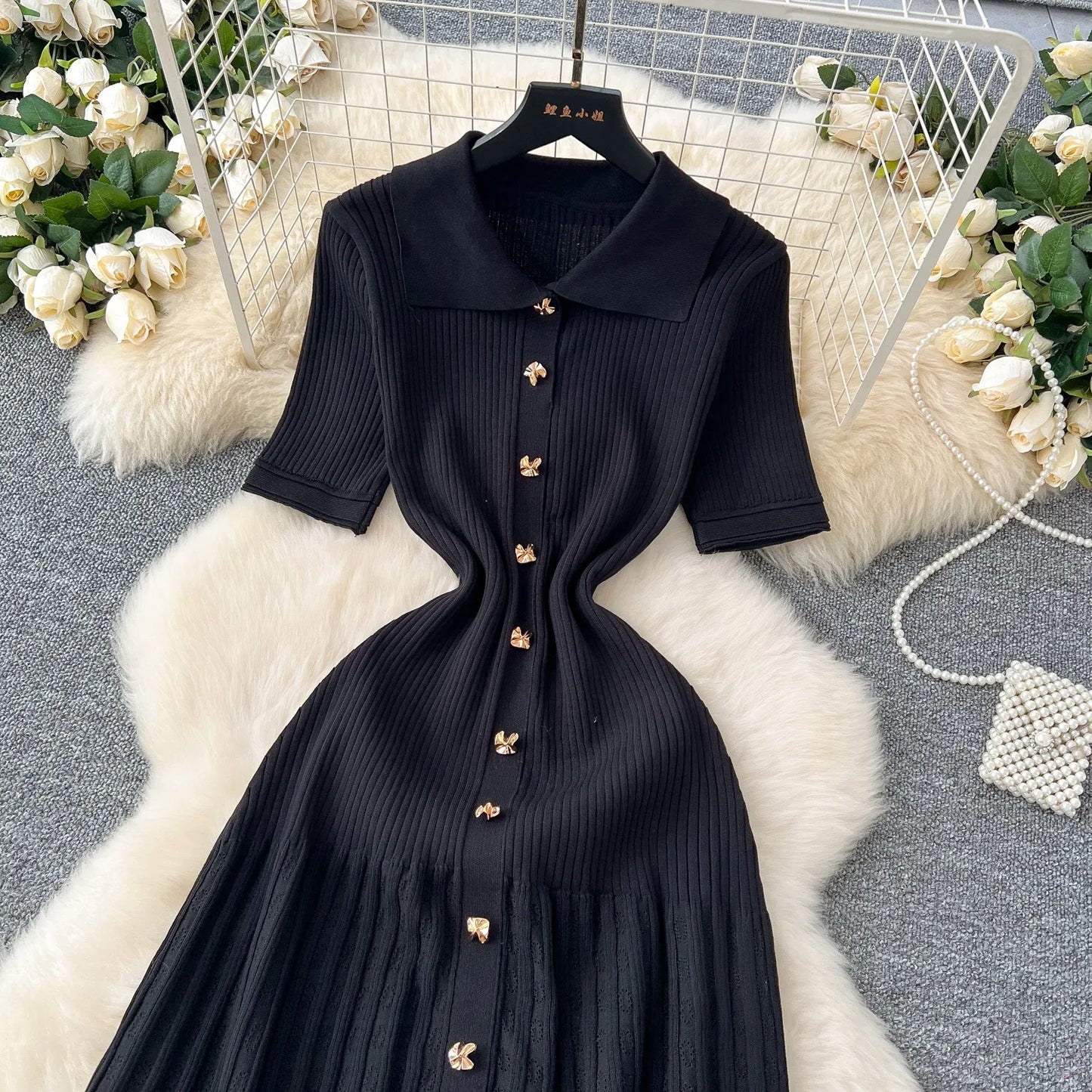Black dress with gold buttons on a hanger against a decorative background with flowers and a rug.
