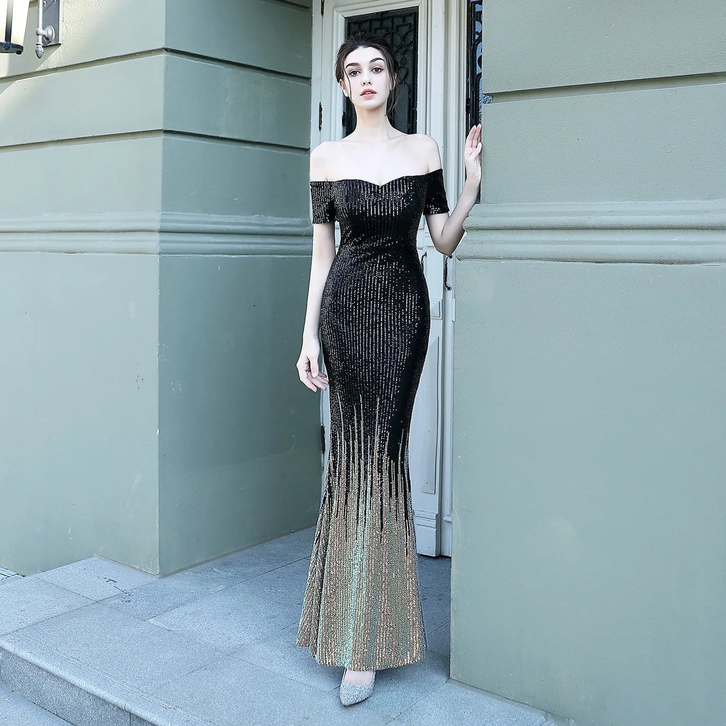 Woman in a black and gold off-shoulder evening gown standing against a light green wall.
