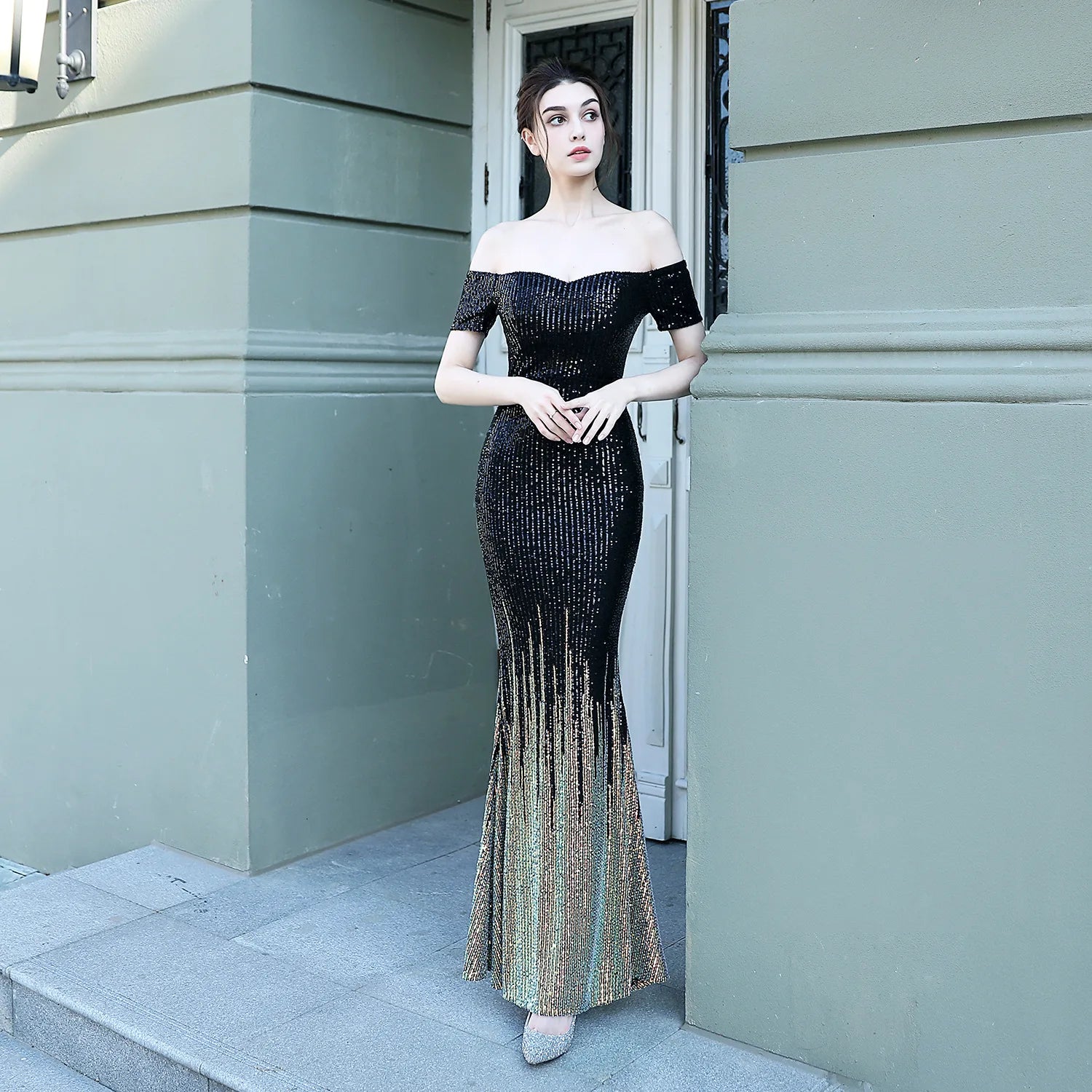 Woman in a black off-shoulder evening gown standing against a light-colored wall.