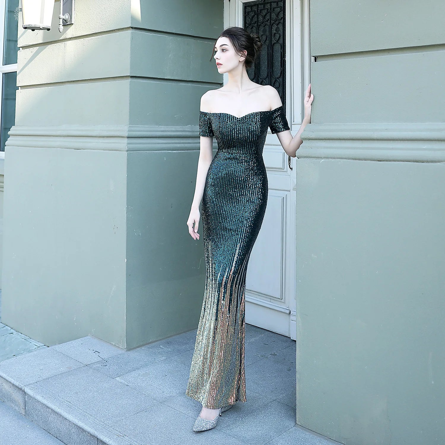 Woman in a black off-shoulder evening gown standing against a light gray building.