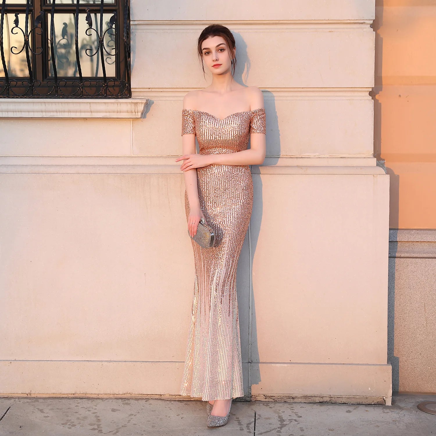 Woman in a gold evening gown standing against a building wall.