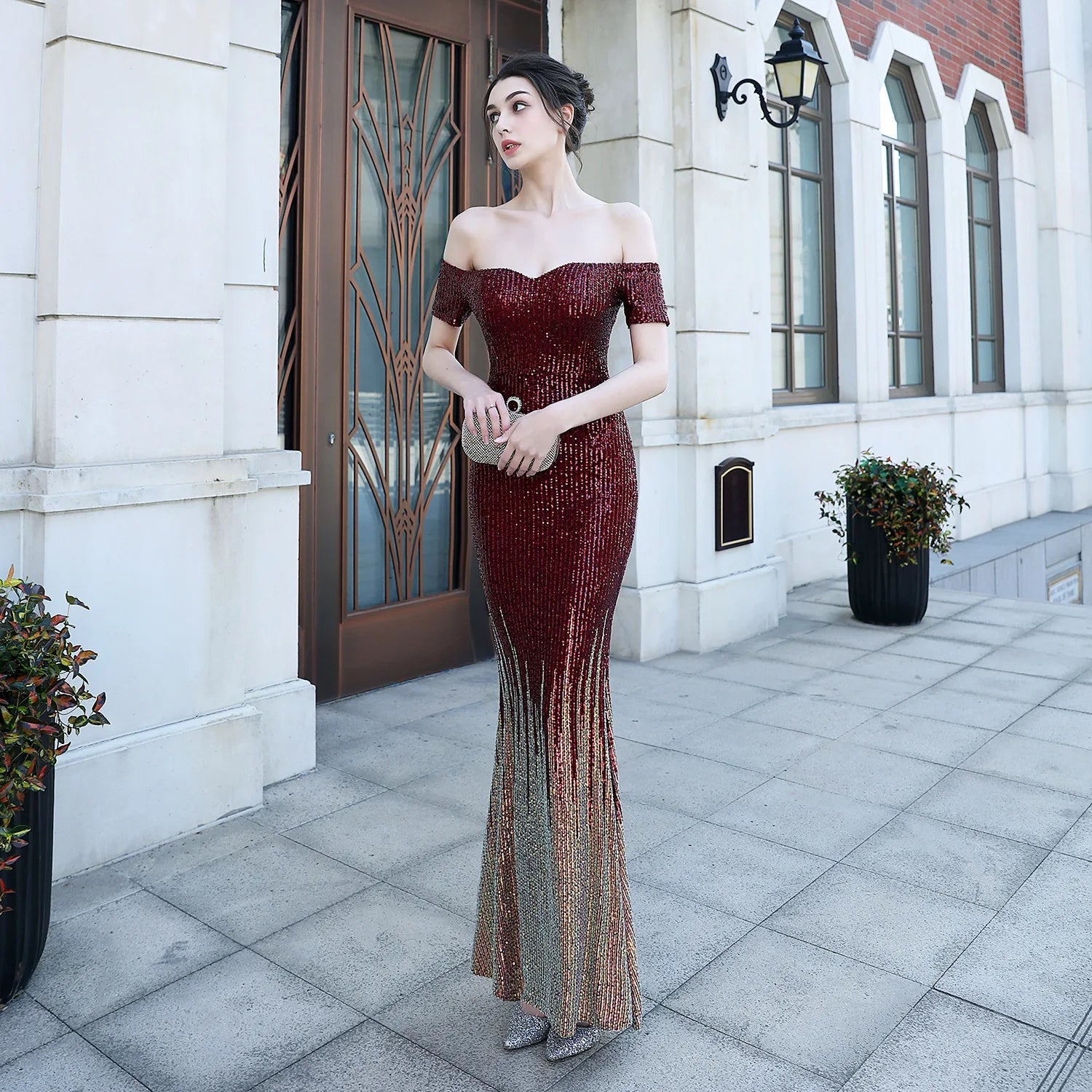 Woman in a burgundy off-shoulder evening gown standing in front of a building entrance.