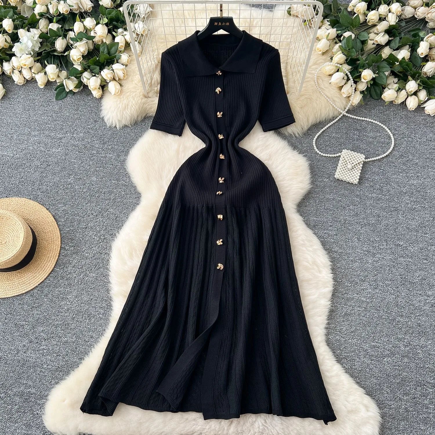 Black dress with gold buttons on a white fur rug surrounded by flowers and a hat.