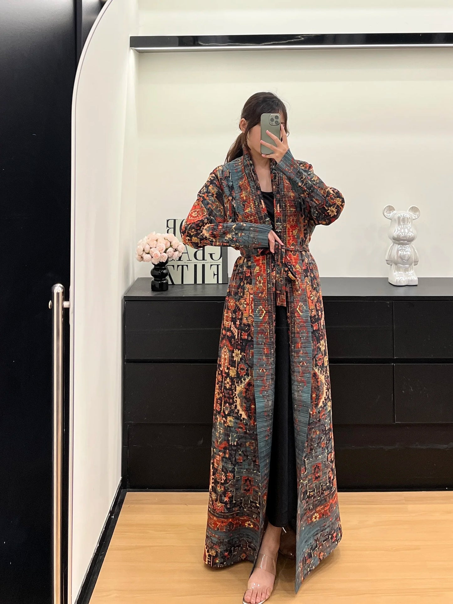 Person wearing a long, patterned coat in a room with a black dresser and decorative items.