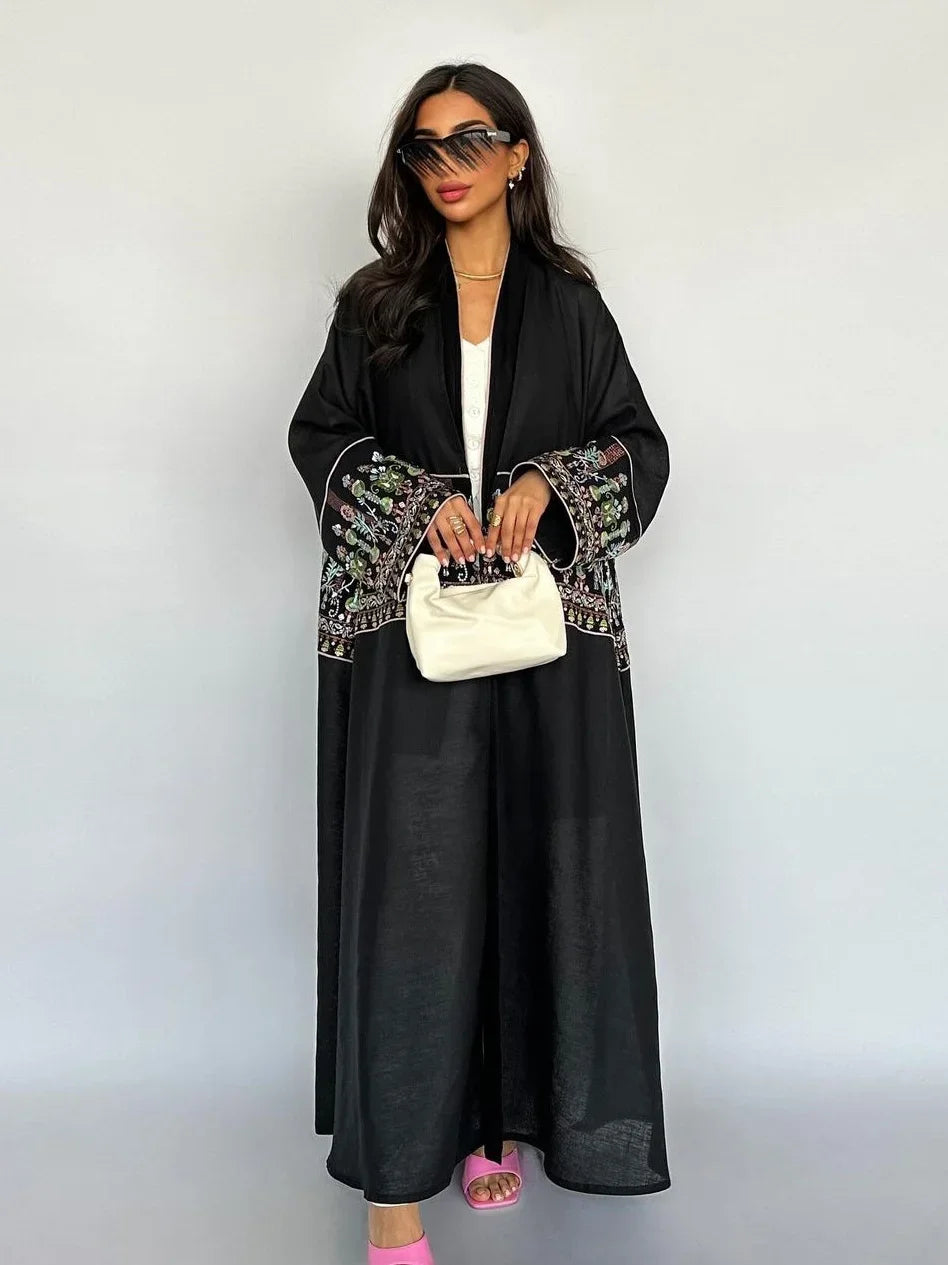 Woman wearing a black outfit with intricate patterns, holding a white handbag against a plain background