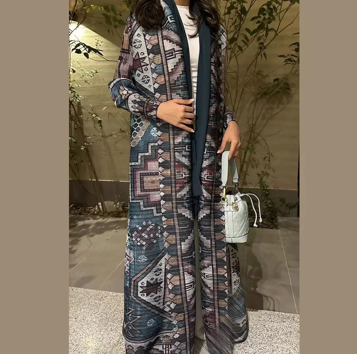Person wearing a long, patterned coat with a white handbag in an outdoor setting.