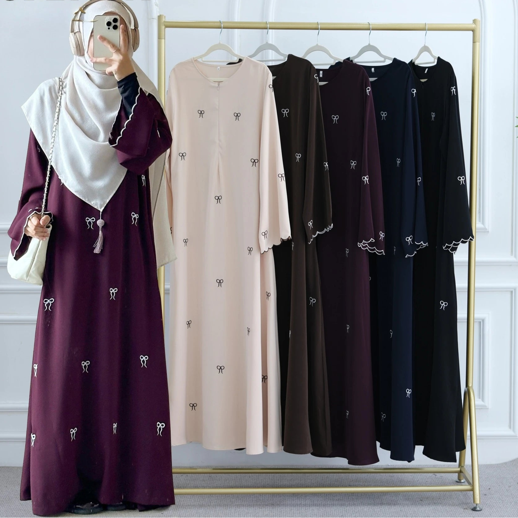 Woman wearing a long dress with a matching hijab, standing next to a rack of similar dresses in various colors.