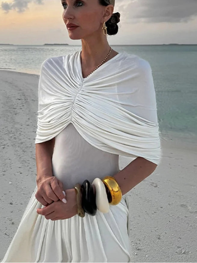 Woman in a white dress with a shawl on a beach, wearing gold jewelry, with 'ZAHM' brand visible.