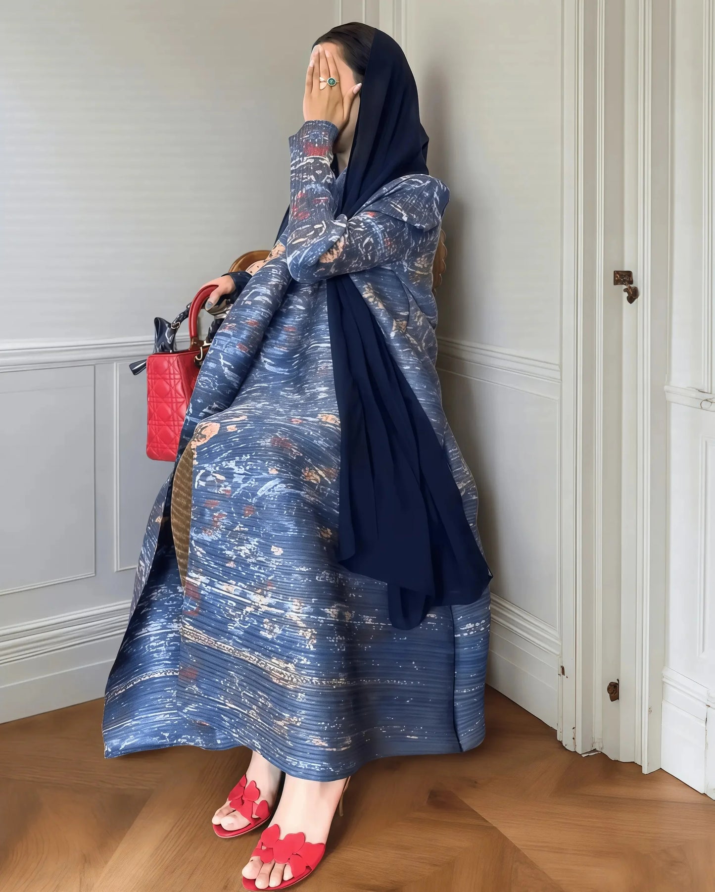 Woman in a blue patterned dress and headscarf standing against a white wall.