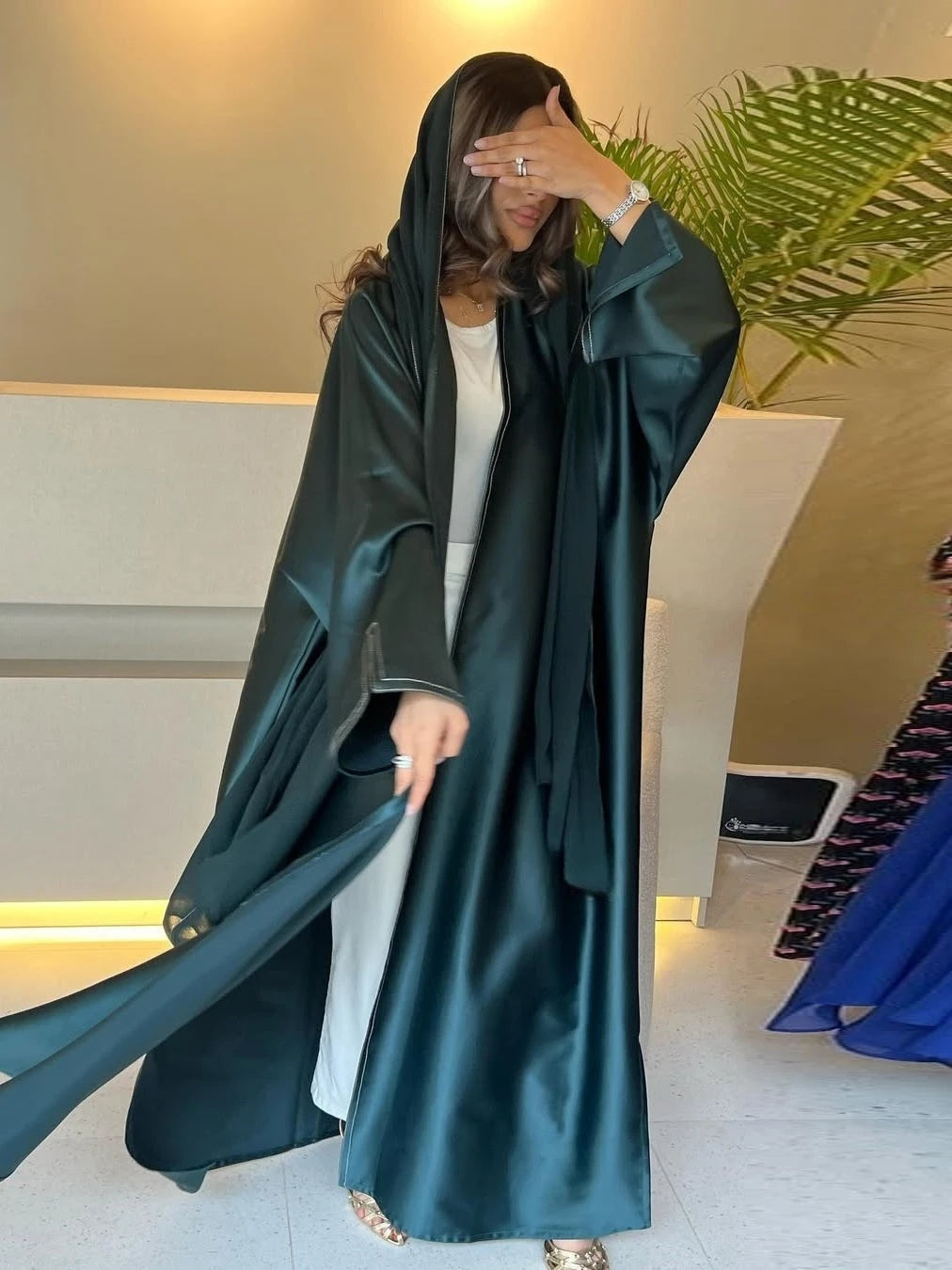 Woman wearing a long dark green robe in an indoor setting with a plant in the background.