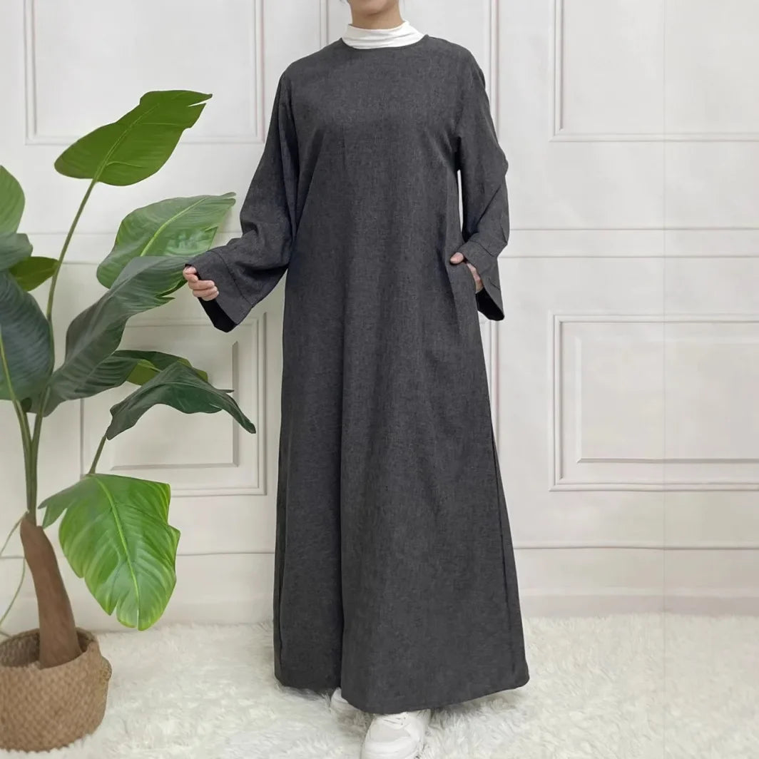 Person wearing a long dark gray dress standing indoors with a plant in the background