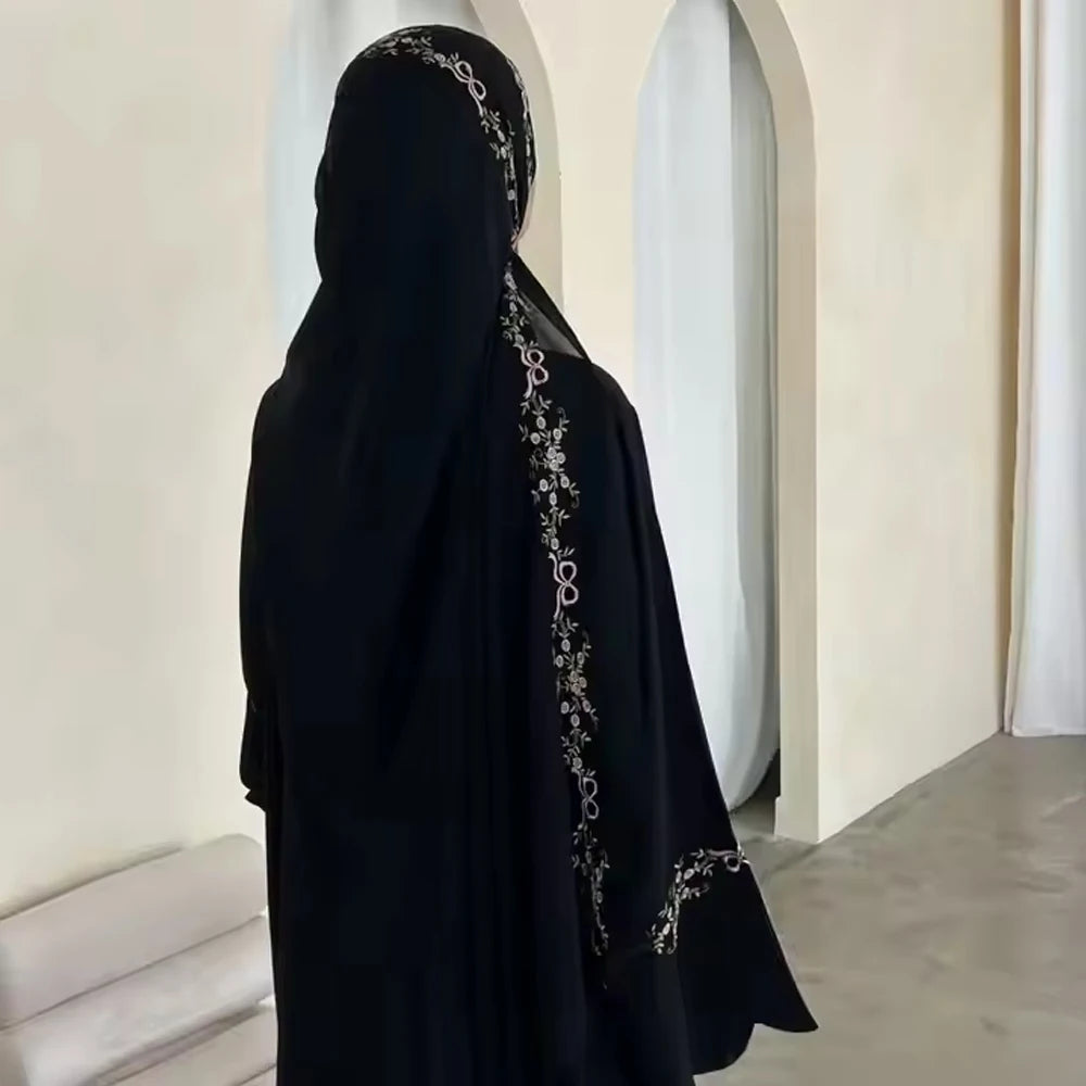 Person wearing a black abaya with intricate silver embroidery in a minimalistic setting.