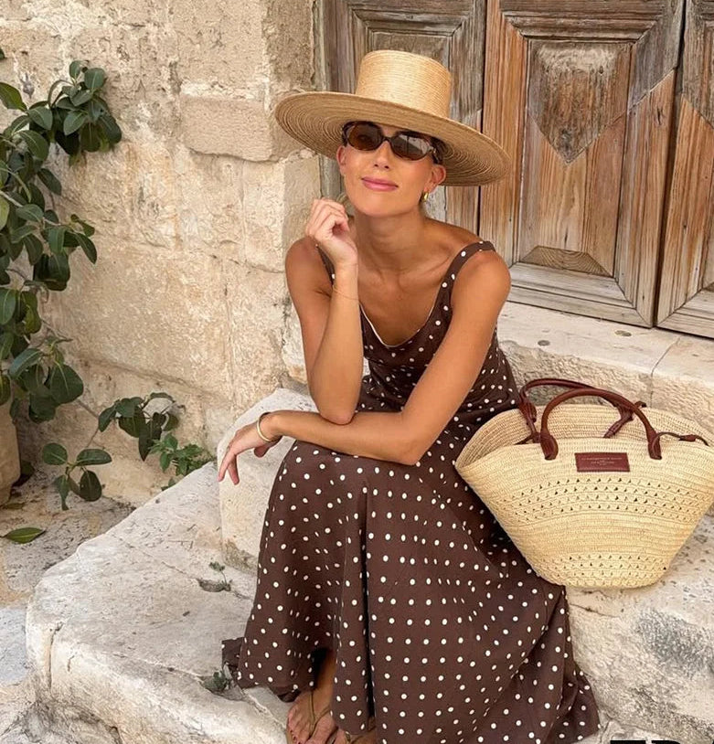 Woman in a brown polka dot dress and wide-brimmed hat sitting on stone steps with a straw bag, against a rustic stone wall.