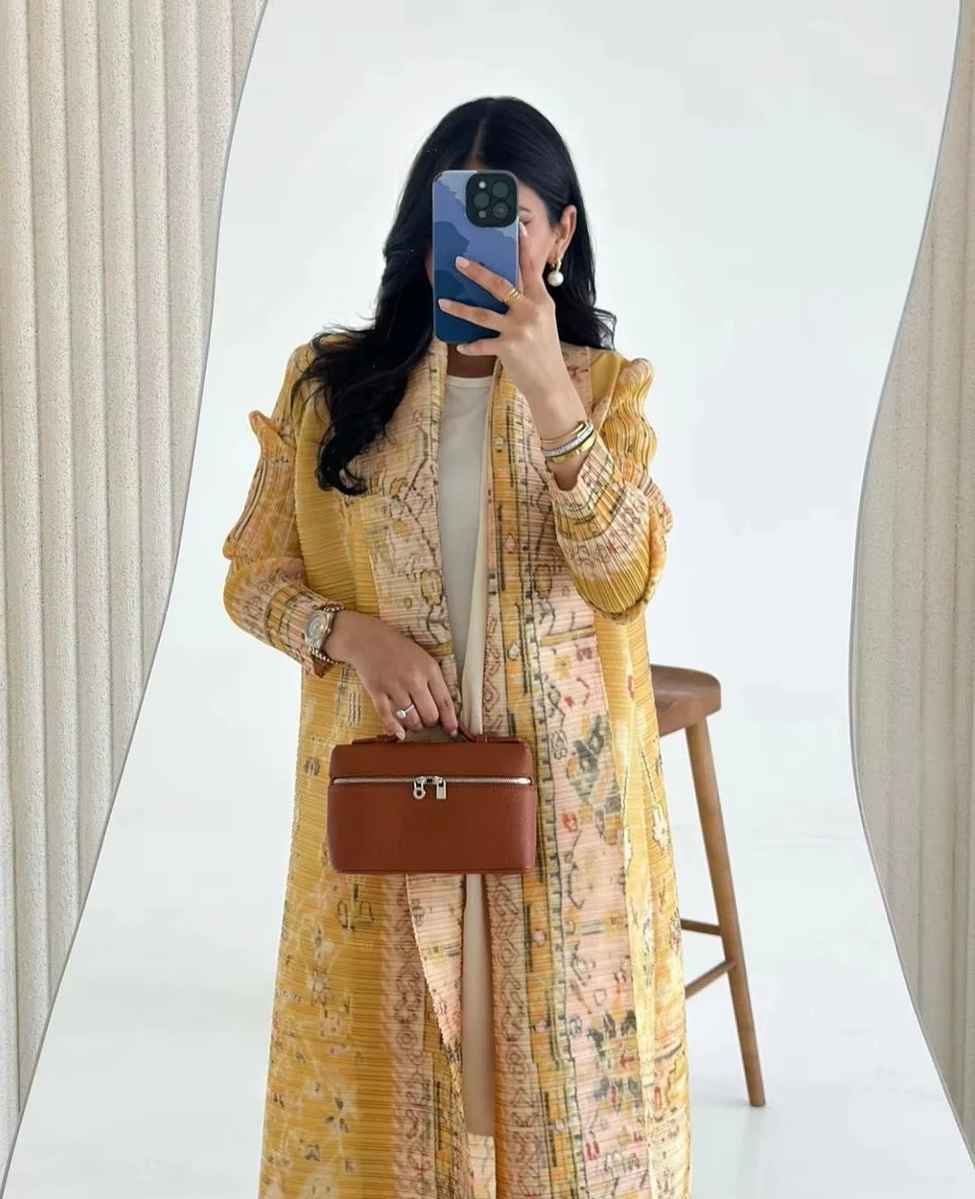 Woman in a yellow floral dress holding a brown handbag in front of a white curtain.