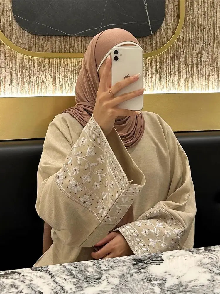 Person wearing a beige hijab and outfit with floral patterns, taking a mirror selfie.