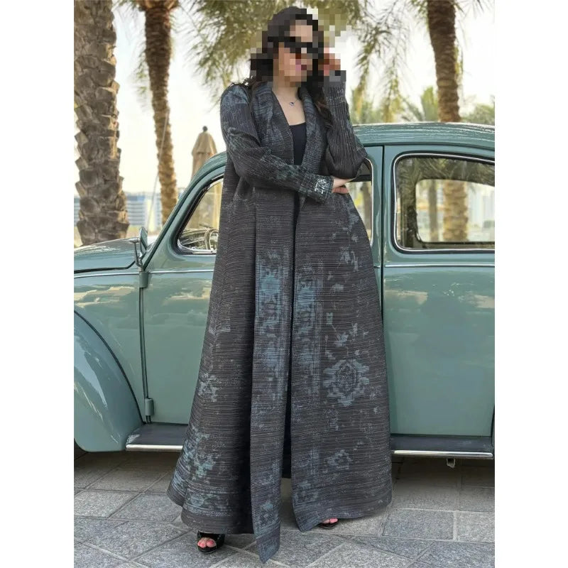 Woman in a long, patterned coat standing next to a vintage car with palm trees in the background.