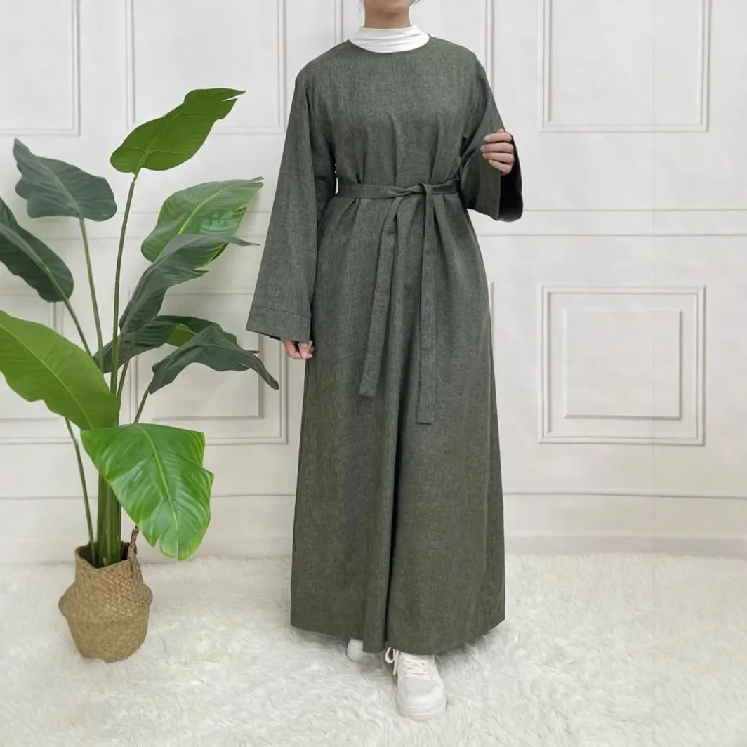 Person wearing a long green dress standing in a room with a plant and white walls.
