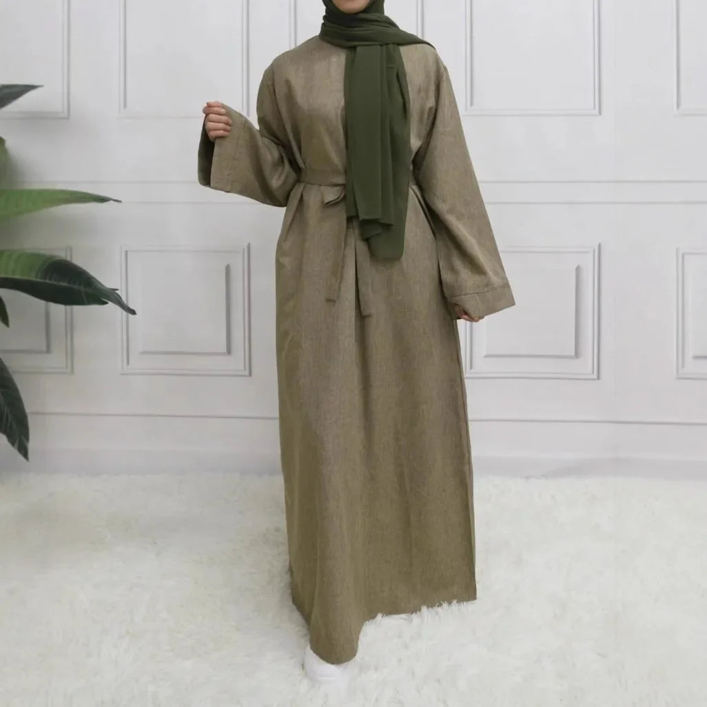 Person wearing a long beige dress with a green hijab in a room with white walls and a plant.