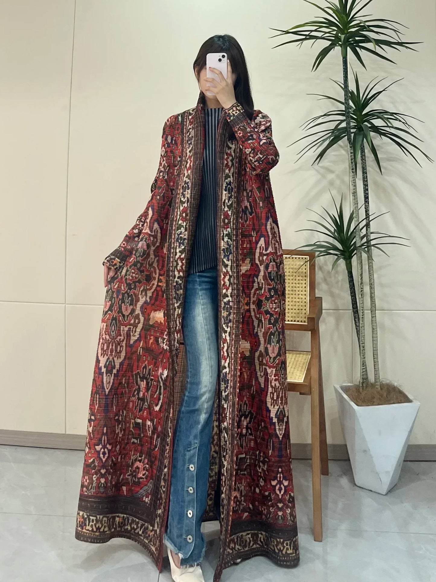 Person wearing a long patterned coat in an indoor setting with a plant and chair.