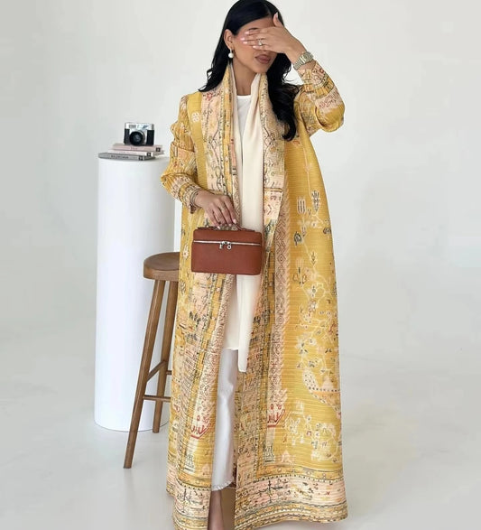 Woman wearing a long yellow patterned coat over a white dress, holding a brown handbag against a white background.