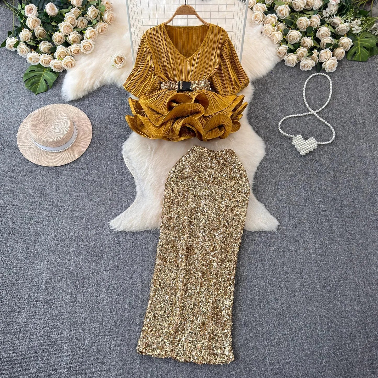 Gold dress with ruffled top and sequin skirt on a mannequin, surrounded by flowers and a hat.