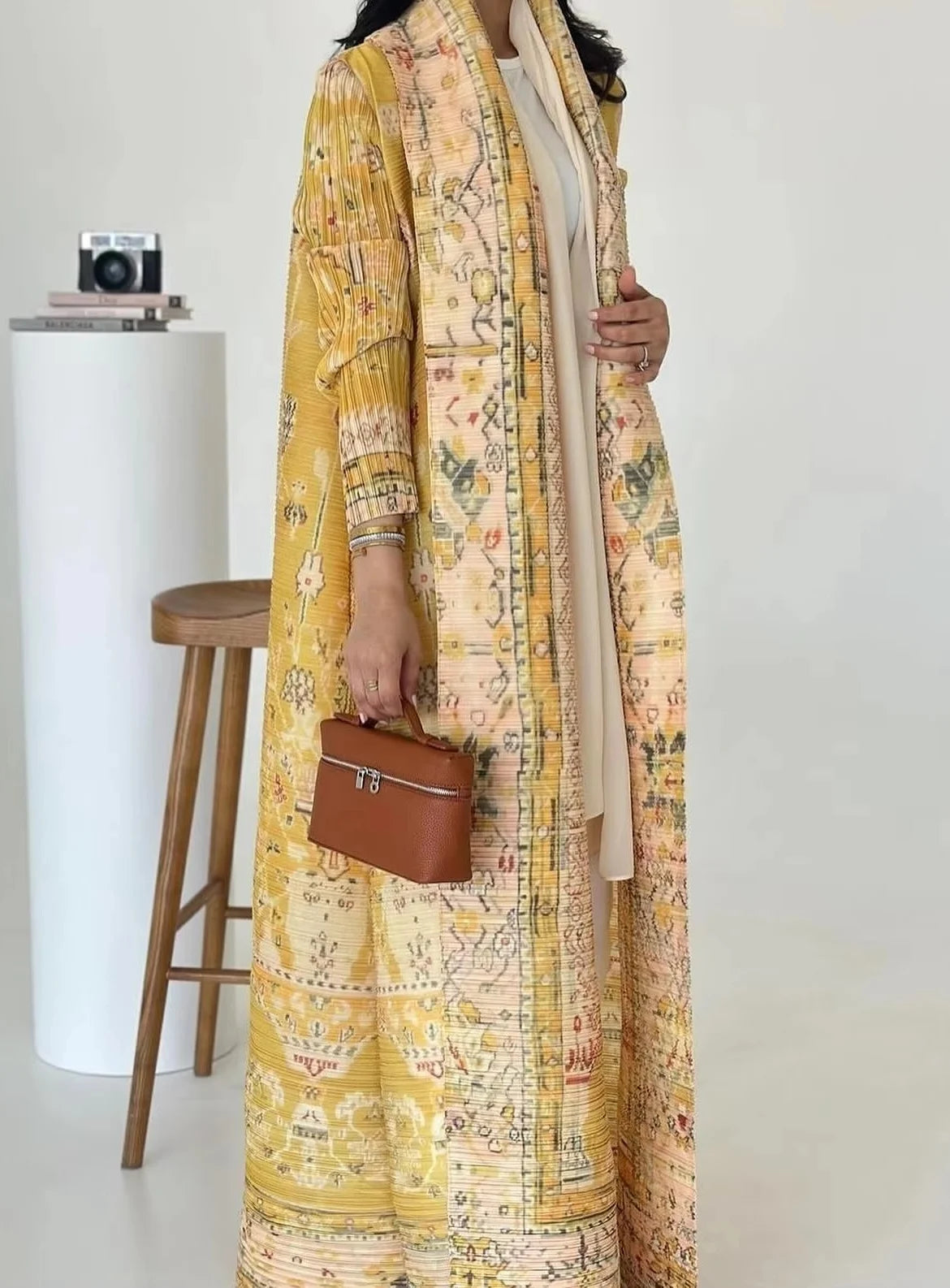 Person wearing a long, patterned yellow coat holding a brown handbag against a white background.
