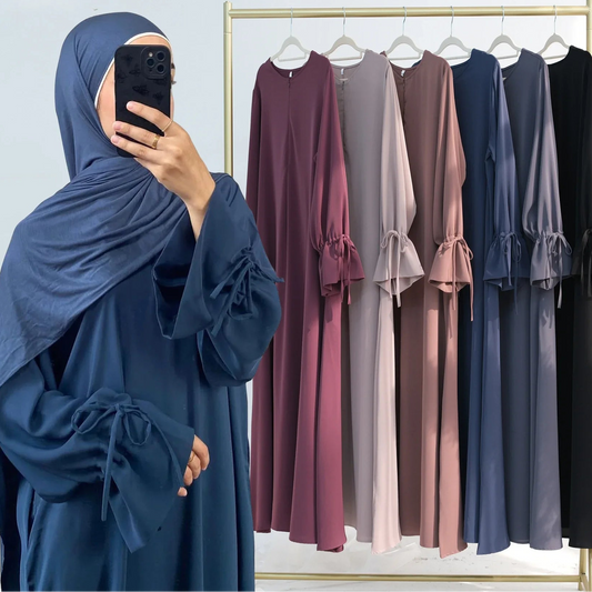 Woman taking a mirror selfie wearing a blue abaya with a rack of various abayas in the background.