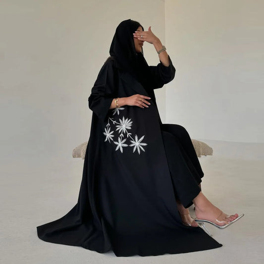Woman wearing a black abaya with white floral embroidery on a plain background