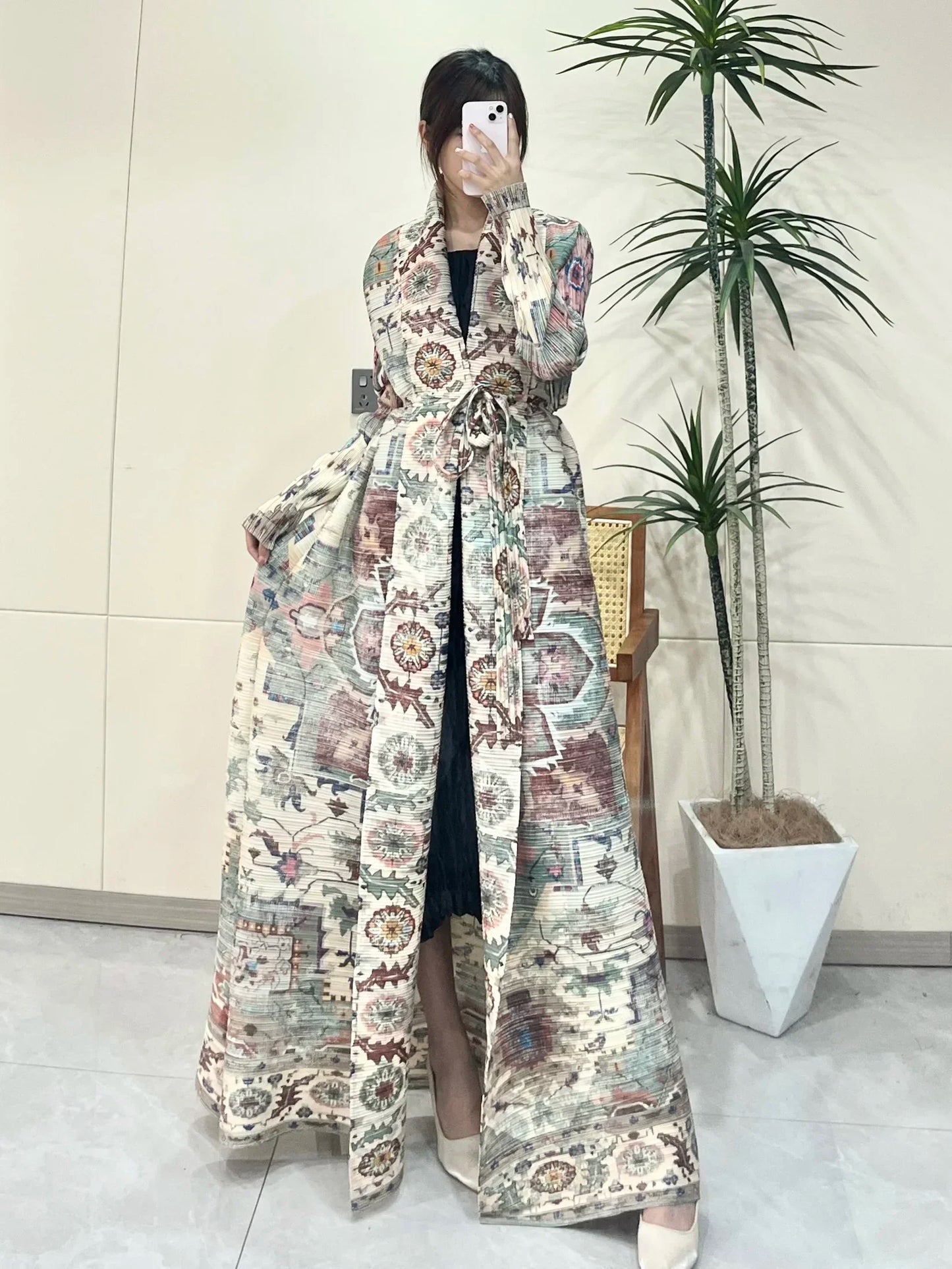 Person wearing a long, patterned coat in an indoor setting with a plant in the background.