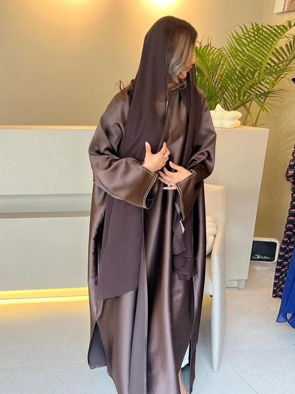 Woman wearing a long, dark brown robe in an indoor setting with a plant and white furniture.