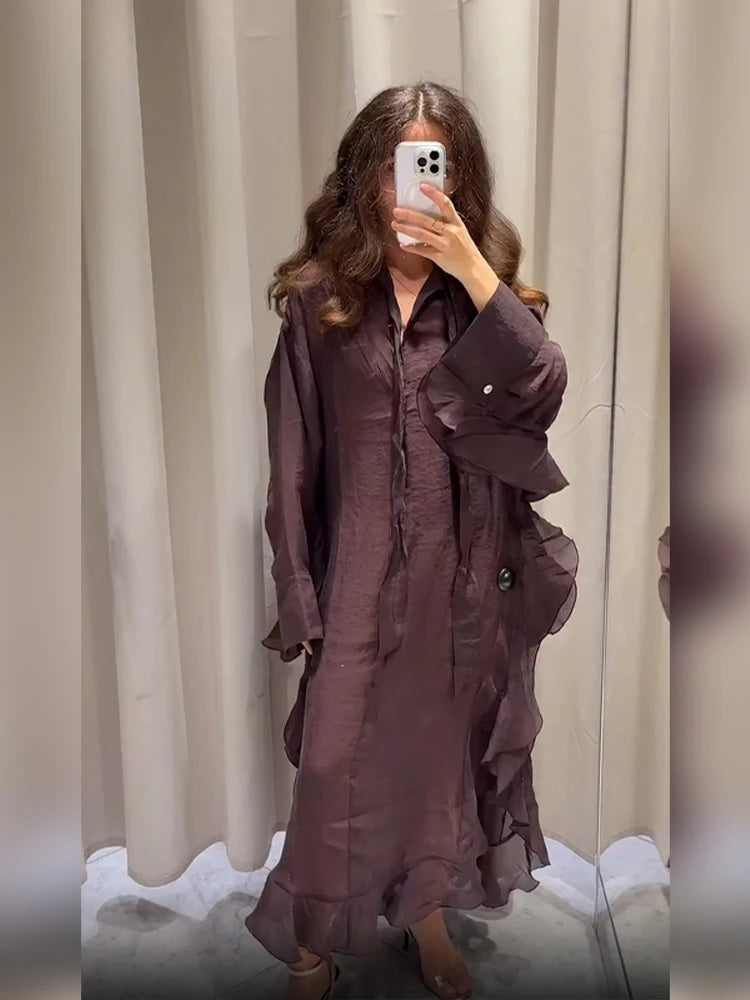 Person wearing a long, dark brown dress standing against a neutral curtain background.
