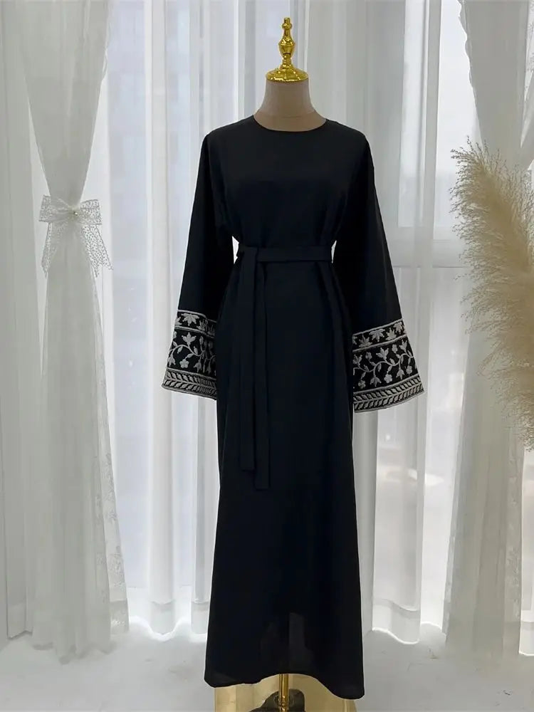 Black dress with white embroidery on long sleeves and waistband on a mannequin against a white curtain background.