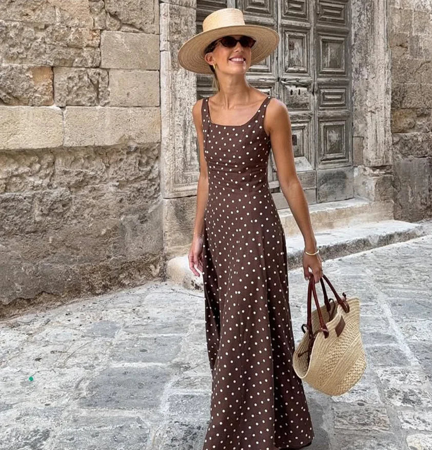 Woman in a polka dot dress and straw hat standing in front of an old stone building, holding a straw bag.