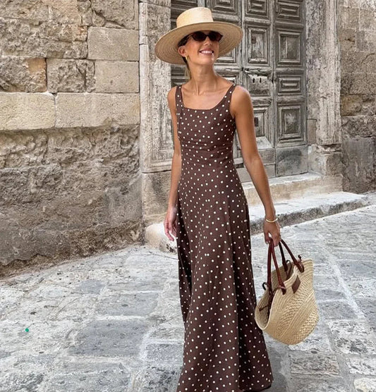 Woman in a polka dot dress and straw hat standing in front of an old stone building, holding a straw bag.