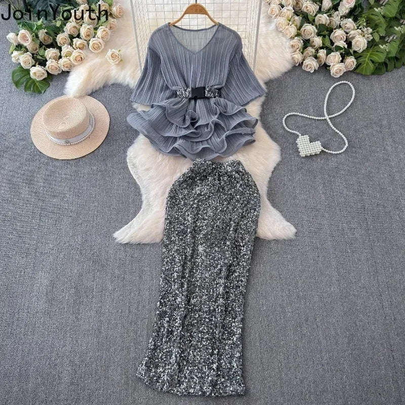 Gray ruffled top and black sequin skirt set on a gray surface with decorative elements.
