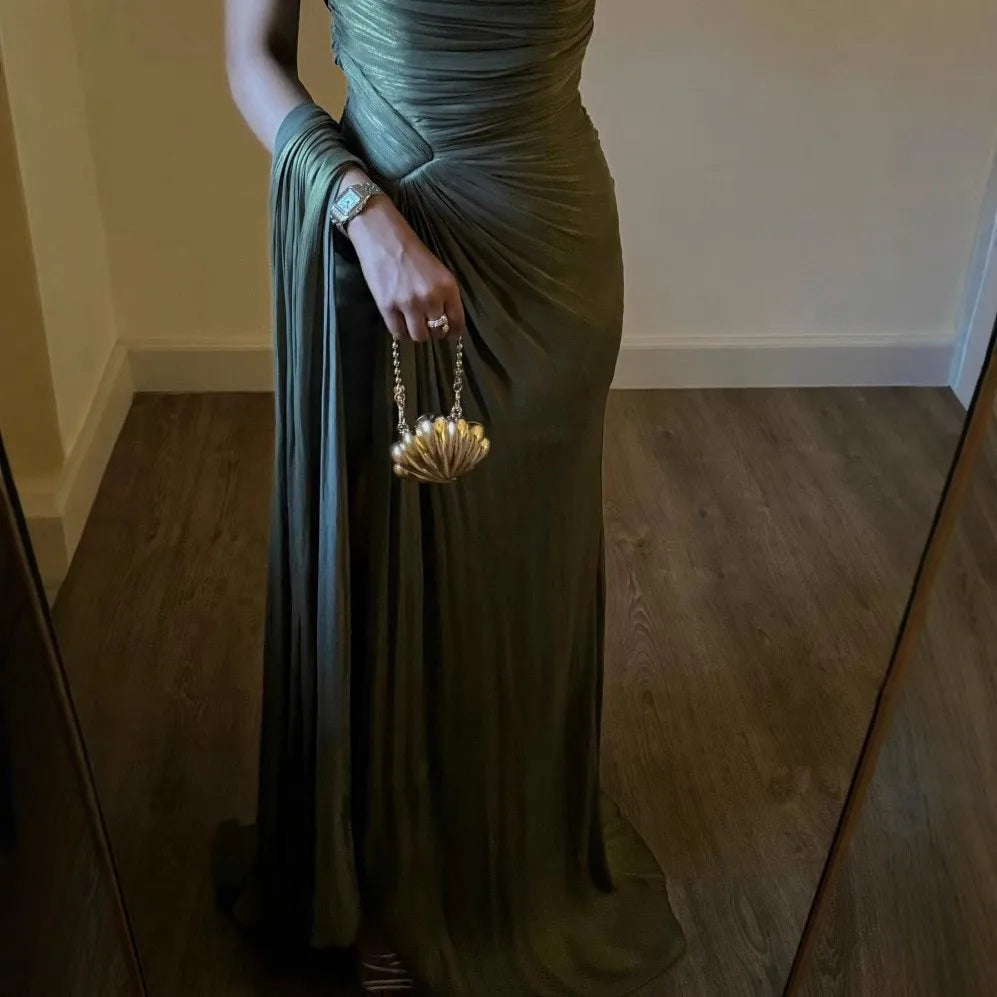 Woman in a green evening gown holding a gold handbag in a room with wooden floor and white walls.