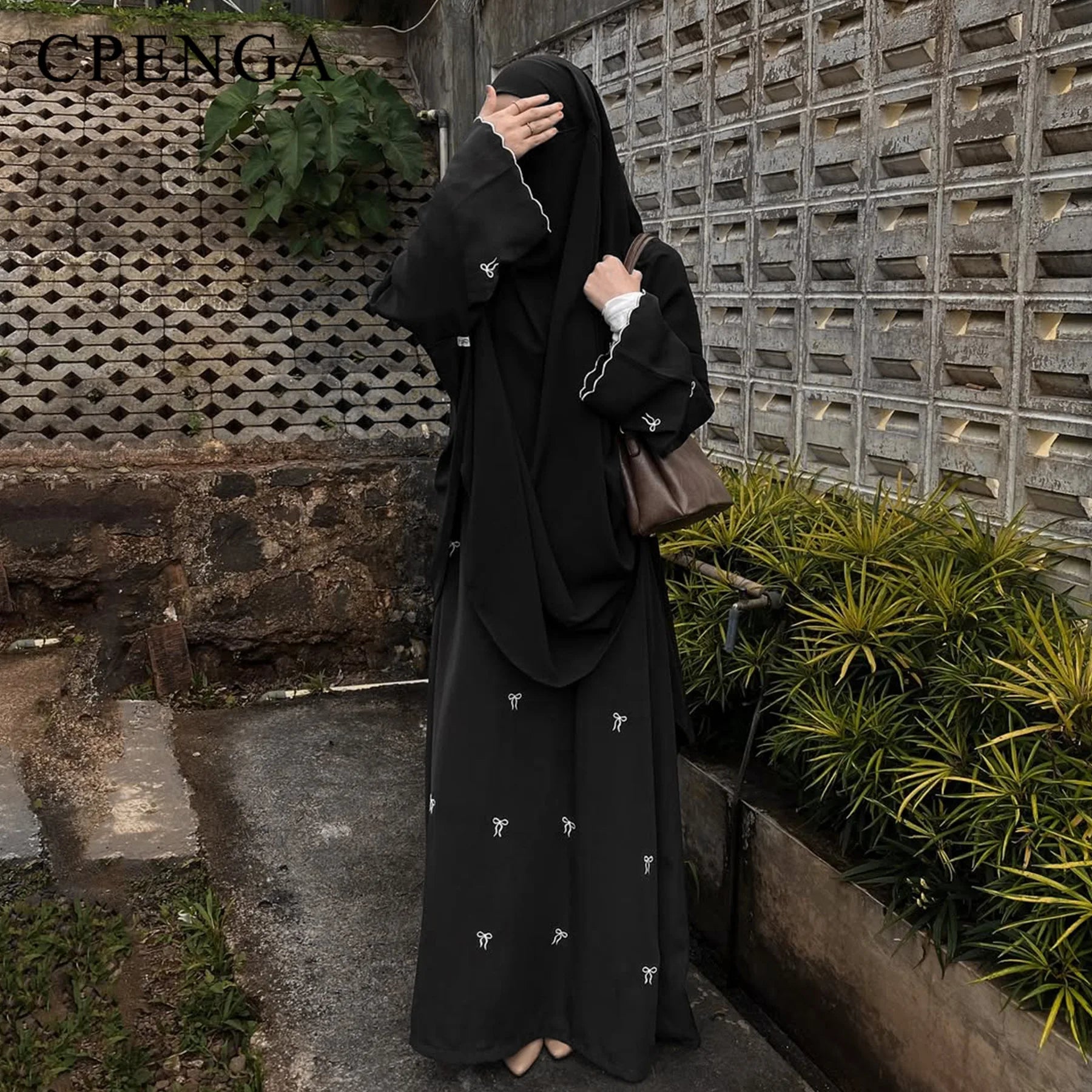 Person wearing a black abaya with white embroidery against a textured wall and plant background.