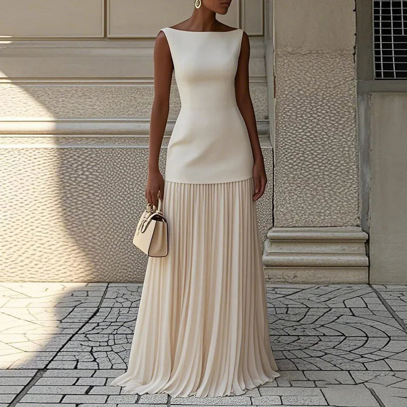 Woman wearing a white sleeveless top and beige pleated skirt standing against a textured wall.