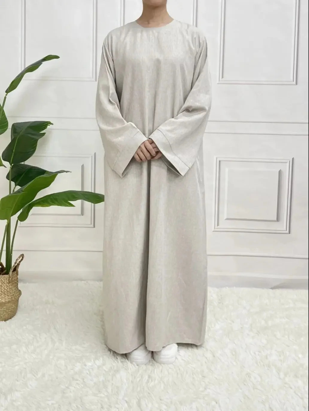 Person wearing a long beige dress standing in a minimalistic room with a plant and white walls.