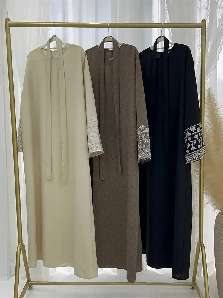 Three abayas in beige, brown, and black hanging on a gold rack against a white curtain background.