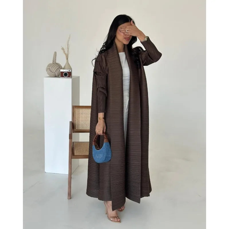 Woman wearing a long brown coat holding a blue handbag in a minimalistic setting.
