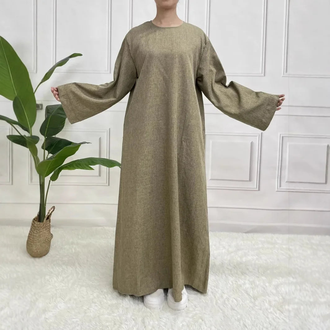 Person wearing a long olive green dress standing in a room with white walls and a plant.