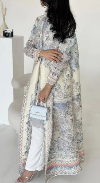 Woman wearing a long, patterned dress over white pants in a minimal indoor setting.