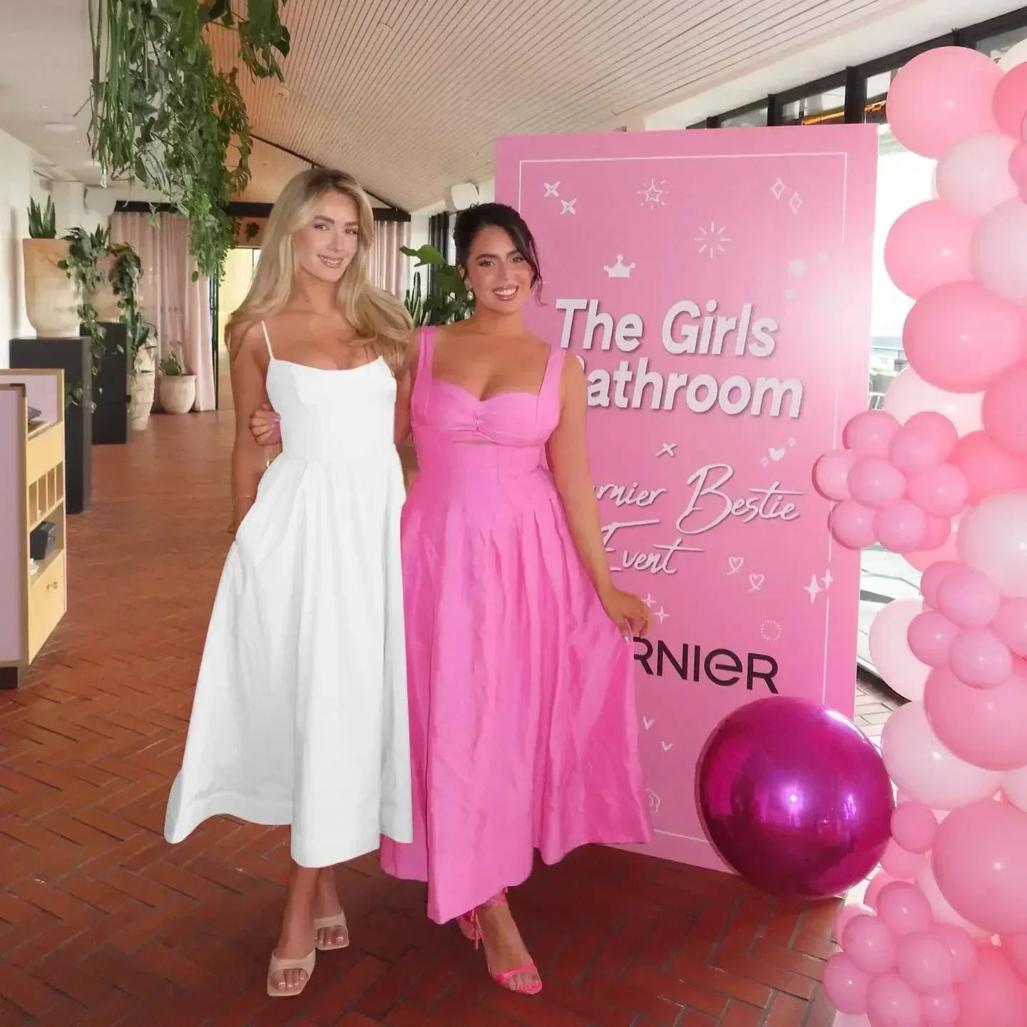 Two women in formal dresses standing next to a pink backdrop with 'The Girls Bathroom' text and balloons.