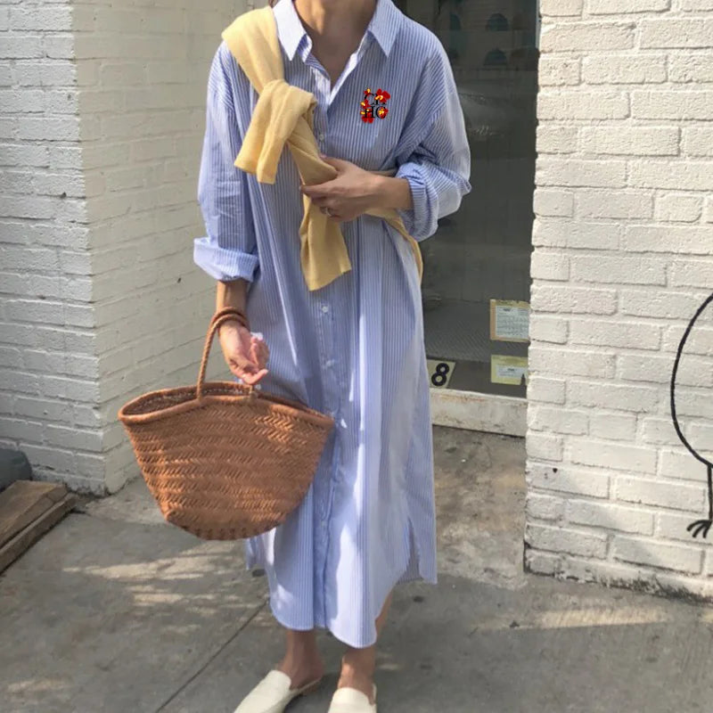 Person wearing a light blue dress with a yellow scarf, holding a woven basket, standing against a brick wall.