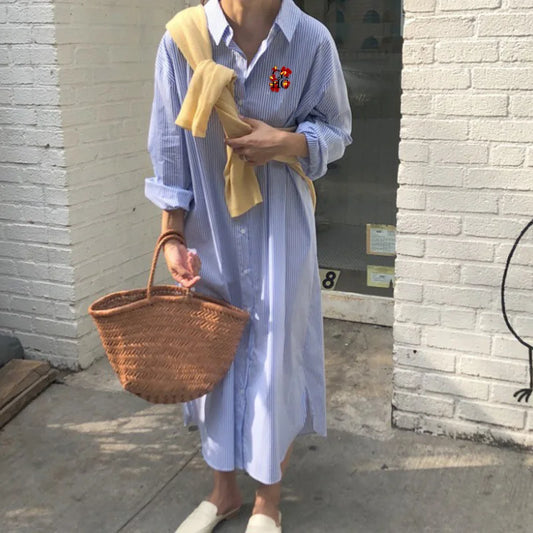 Person wearing a light blue dress with a yellow scarf, holding a woven basket, standing against a brick wall.