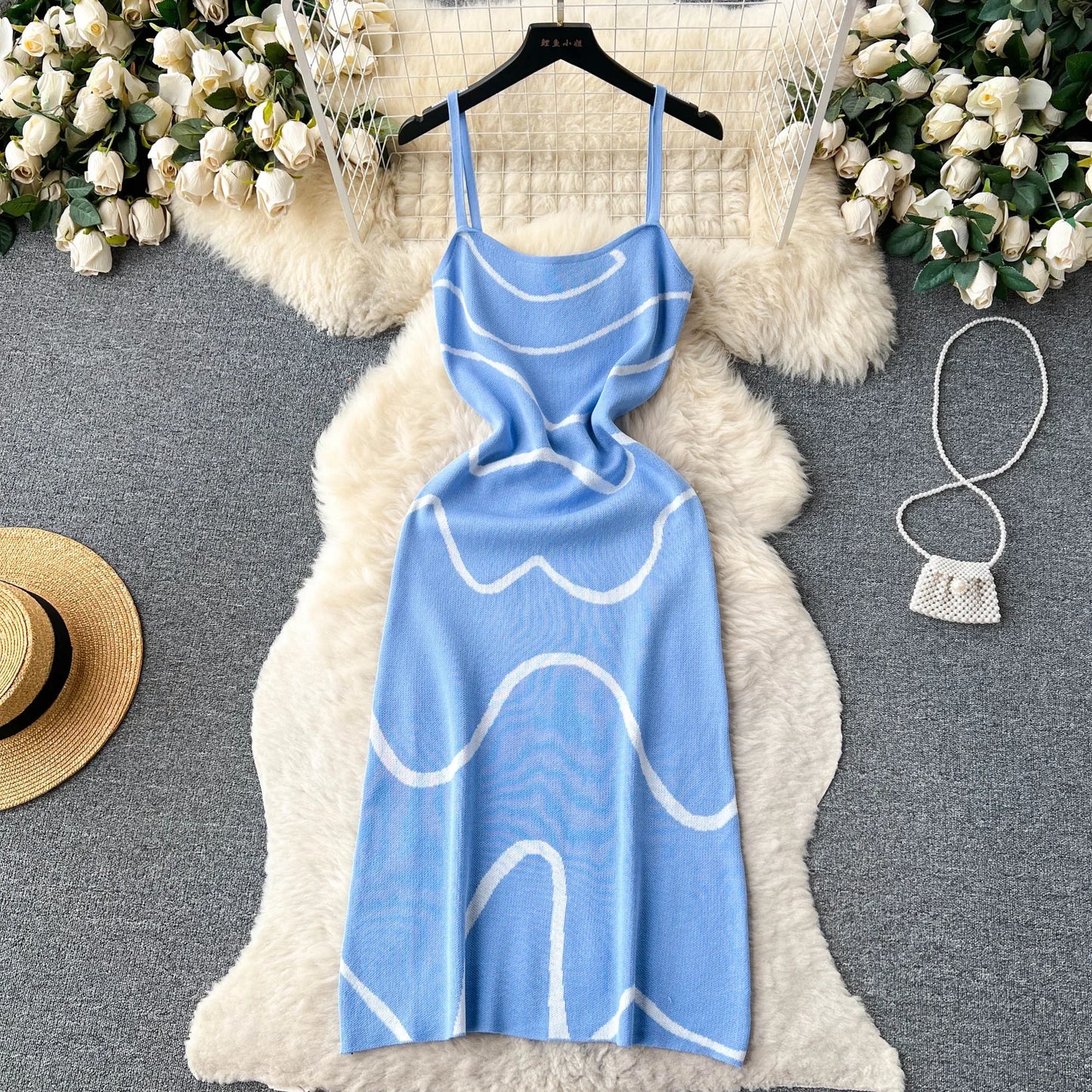 Blue dress with white patterns on a hanger against a decorative background with flowers and a hat.